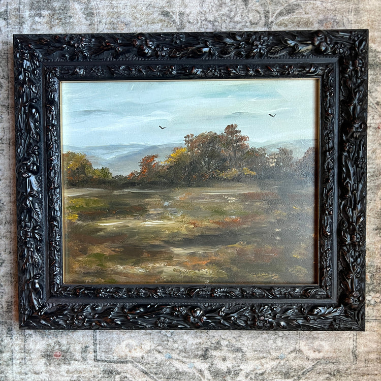 A framed landscape oil painting featuring a misty field, trees, and a pale sky, housed in an ornate carved black wooden frame and displayed against a textured, distressed gray wall.