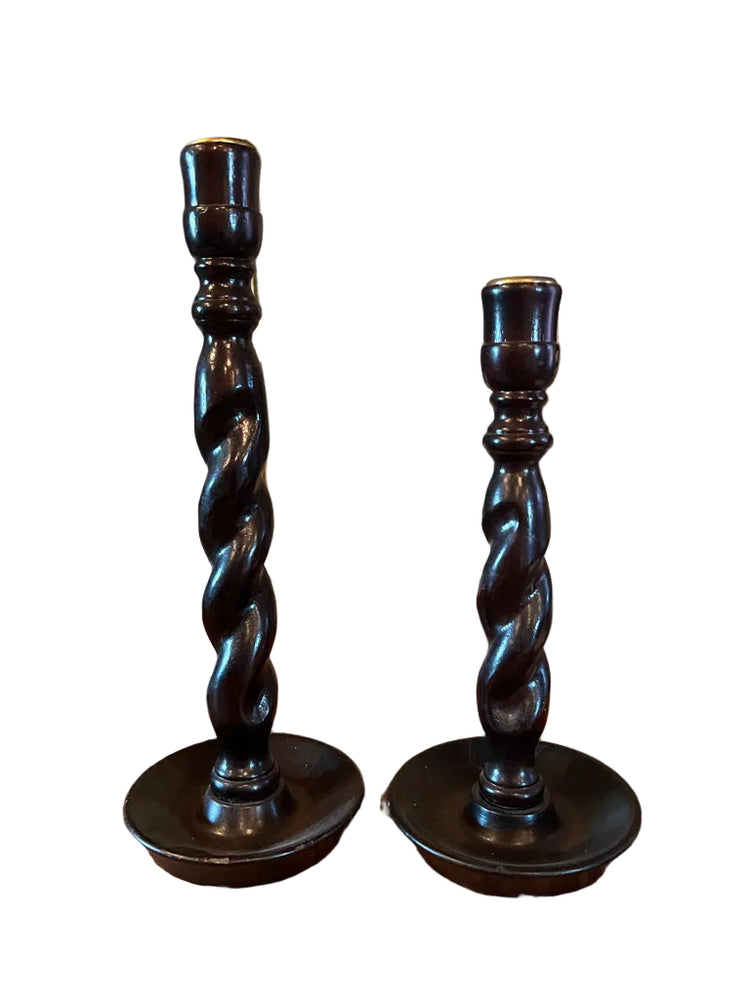 Two twisted wooden candlesticks on a wooden surface with framed paintings in the background.