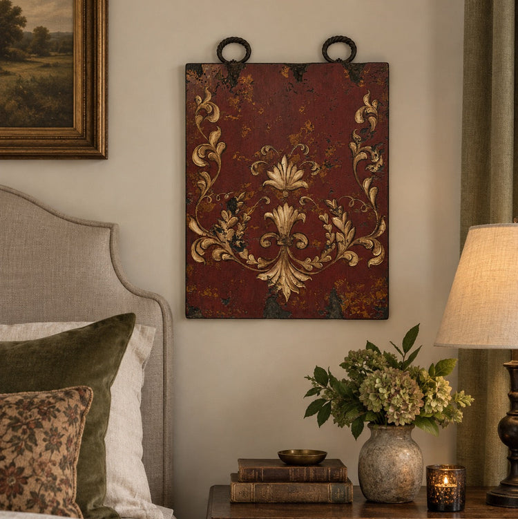 A rectangular wall hanging with a distressed dark red background and ornate gold damask-style scrollwork, displayed above a bedside table in a styled bedroom setting.