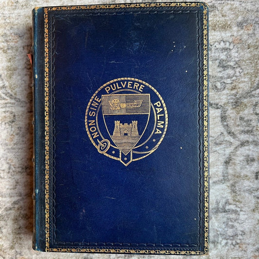 A vintage dark blue leather-bound book or notebook with gold foil detailing and a central crest, placed on a textured gray stone surface.