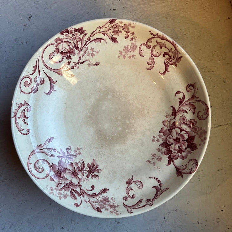 A round ceramic plate with a white base and a dusty rose floral scroll pattern, photographed from above on a gray textured surface.