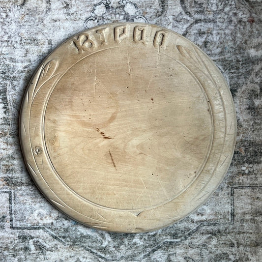 A round, light-colored wooden serving board with carved Hebrew lettering at the top edge, displayed on a distressed, patterned gray rug.
