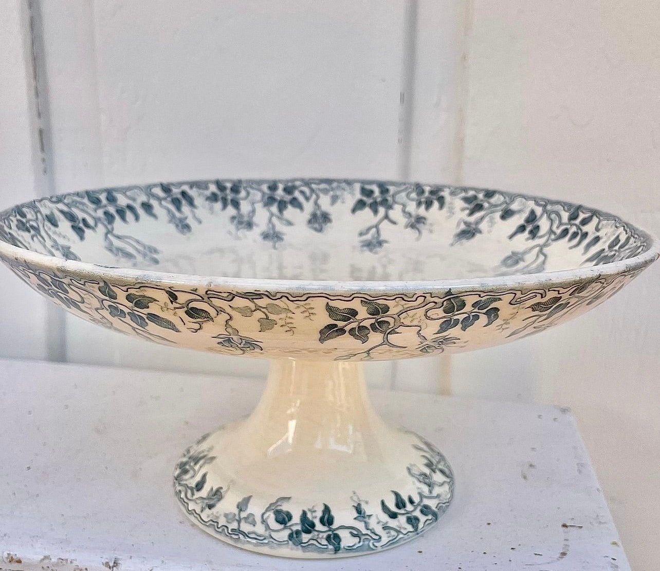A vintage ceramic pedestal plate with a cream-colored glaze and dark blue floral and leaf patterns, displayed on a white surface against a white paneled background.