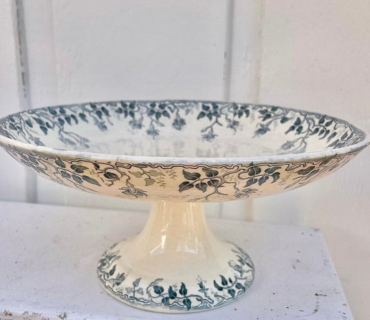 A vintage ceramic pedestal plate with a cream-colored glaze and dark blue floral and leaf patterns, displayed on a white surface against a white paneled background.
