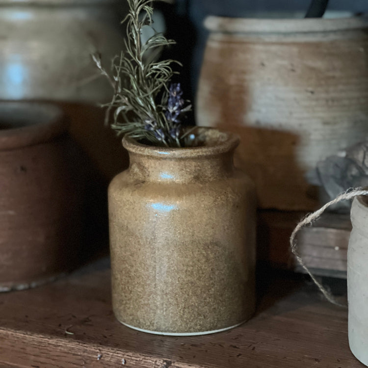 A small handmade ceramic jar with a speckled brown and beige glaze, holding dried green and purple herbs, displayed on a rustic wooden shelf.
