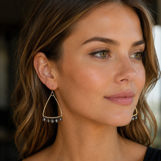 A close-up of a woman wearing gold triangle dangle earrings with small dark bead accents, shown from the chest up against a softly blurred background.