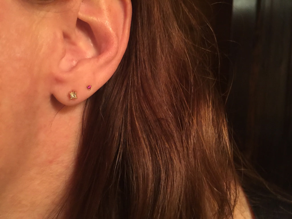 Close-up of a person's ear showing two small stud earrings, one gold and one pink, with brown hair in the foreground.