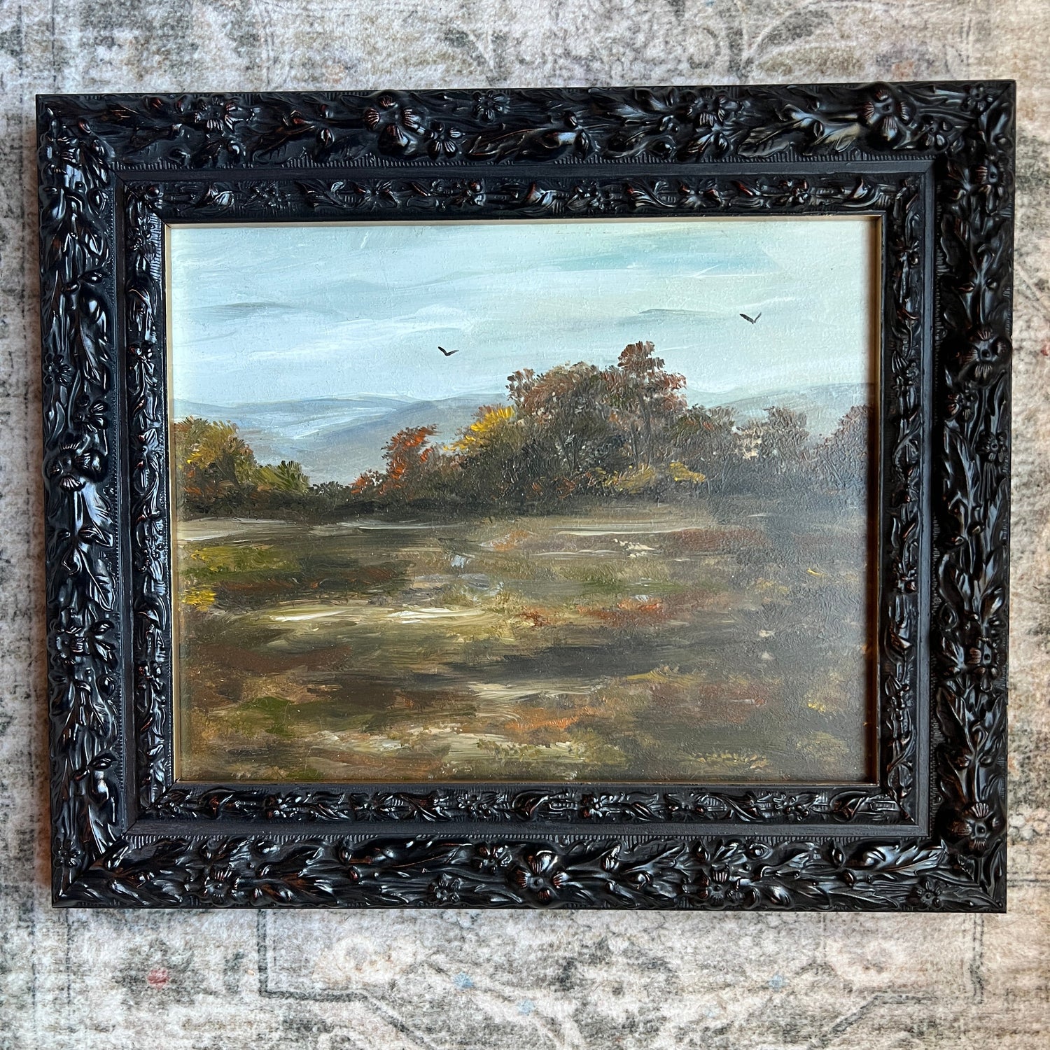 A framed landscape oil painting featuring a misty field, trees, and a pale sky, housed in an ornate carved black wooden frame and displayed against a textured, distressed gray wall.