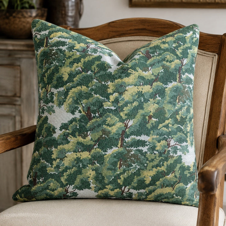 A square throw pillow with a green and yellow forest tree pattern, resting on a beige upholstered wooden armchair in a cozy room setting.