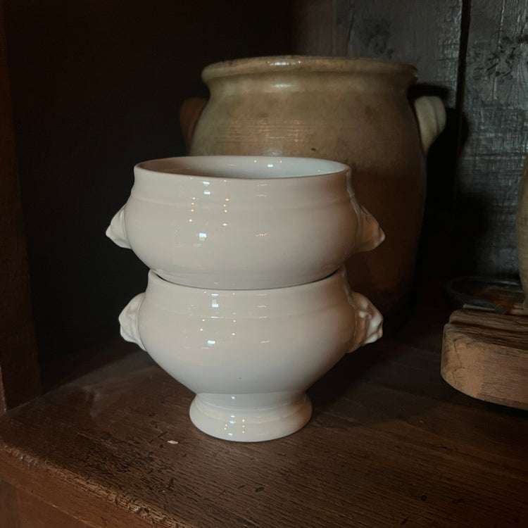 A pair of stacked white ceramic cups with side handles, displayed on a rustic wooden shelf with a larger clay pot in the background.