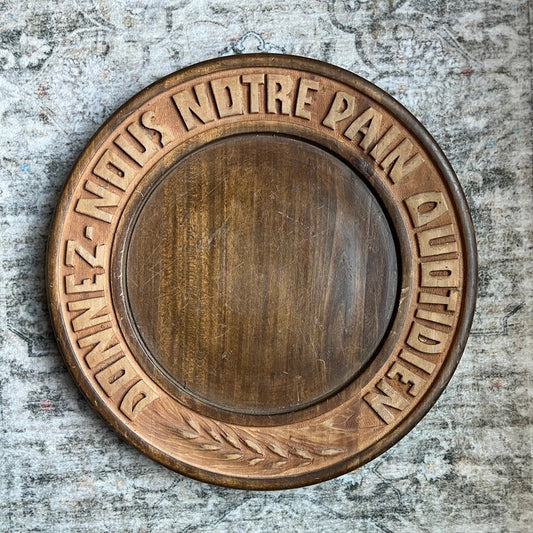 A round wooden plaque with a carved border featuring the French text 'DONNEZ-NOUS NOTRE PAIN OU VDIDIDLEN' and a leaf motif, mounted on a textured gray plaster wall.