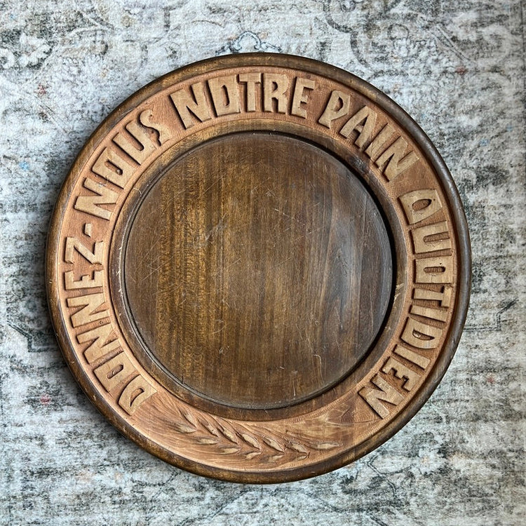 A round wooden plaque with a carved border featuring the French text 'DONNEZ-NOUS NOTRE PAIN OU VDIDIDLEN' and a leaf motif, mounted on a textured gray plaster wall.