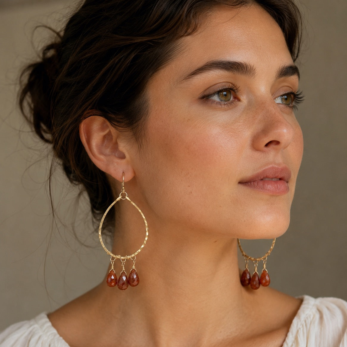 A close-up of a woman wearing large gold-tone hoop earrings with three dangling amber-brown teardrop beads on each earring, paired with a white top against a neutral beige background.