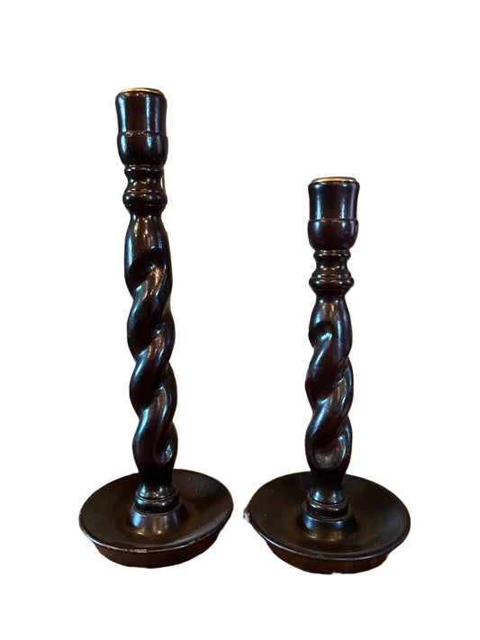 Two twisted wooden candlesticks on a wooden surface with framed paintings in the background.