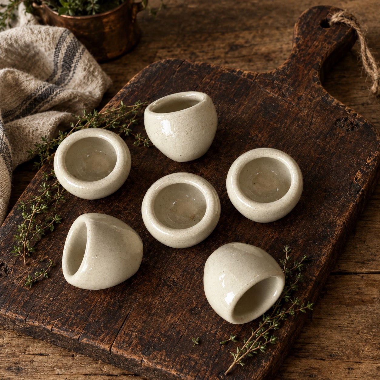 Five small matte ceramic cups arranged on a rustic dark wooden cutting board with sprigs of herbs and a folded cloth nearby.