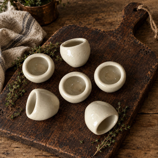 Five small matte ceramic cups arranged on a rustic dark wooden cutting board with sprigs of herbs and a folded cloth nearby.