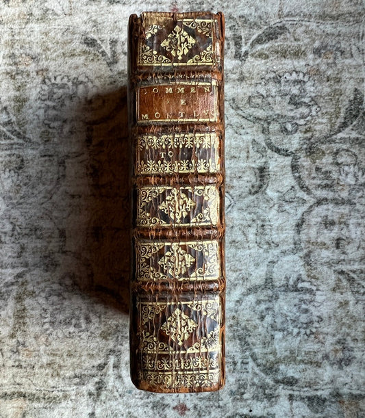 An antique leather-bound book standing upright on a textured, distressed gray surface. The book's spine features ornate gold gilding and tooling on a dark brown leather base, with the title 'GOMMEN NO.