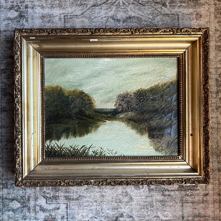 An oil landscape painting of a river with trees on both banks, housed in an ornate gold frame and displayed on a textured, distressed gray wall.