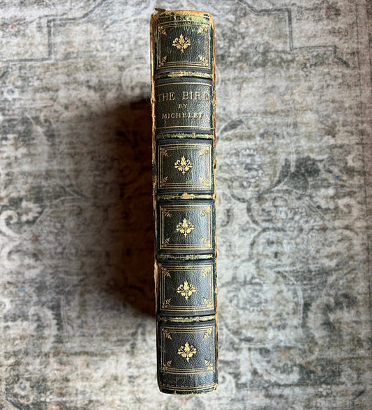 An antique leather-bound book standing upright on its spine, featuring dark green leather with gold tooling and lettering, set against a textured gray background.