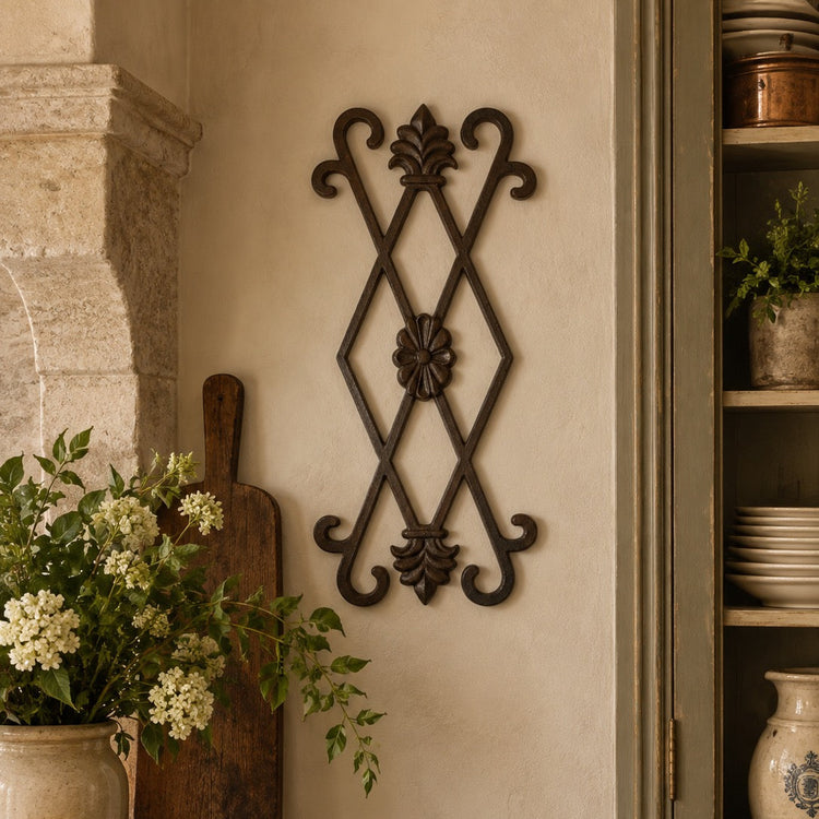 A dark metal decorative wall hanging with an ornate diamond lattice design is mounted on a beige plaster wall in a rustic kitchen setting, flanked by a stone column, a bowl of lemons, and a green cabinet.