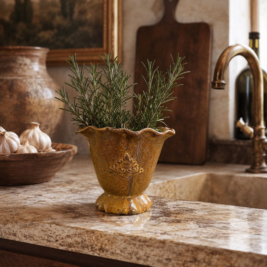 A rustic yellow-glazed ceramic planter with scalloped edges and embossed detailing, filled with green rosemary sprigs, set on a granite kitchen countertop near a sink and brass faucet.