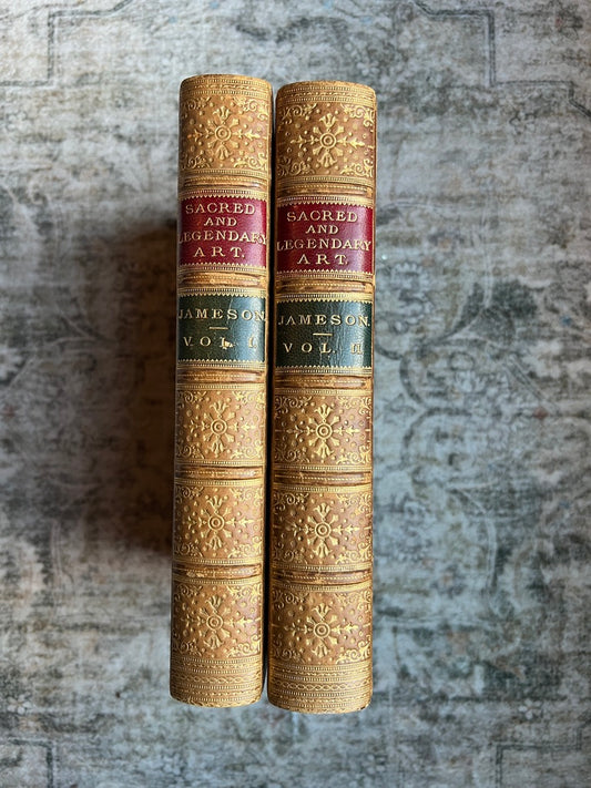 Two antique leather-bound books standing upright side by side on a textured gray stone surface. The spines are gold-toned with ornate floral embossing and red bands displaying the title 'Sacred and Legendary Art' and 'Jameson Vol.