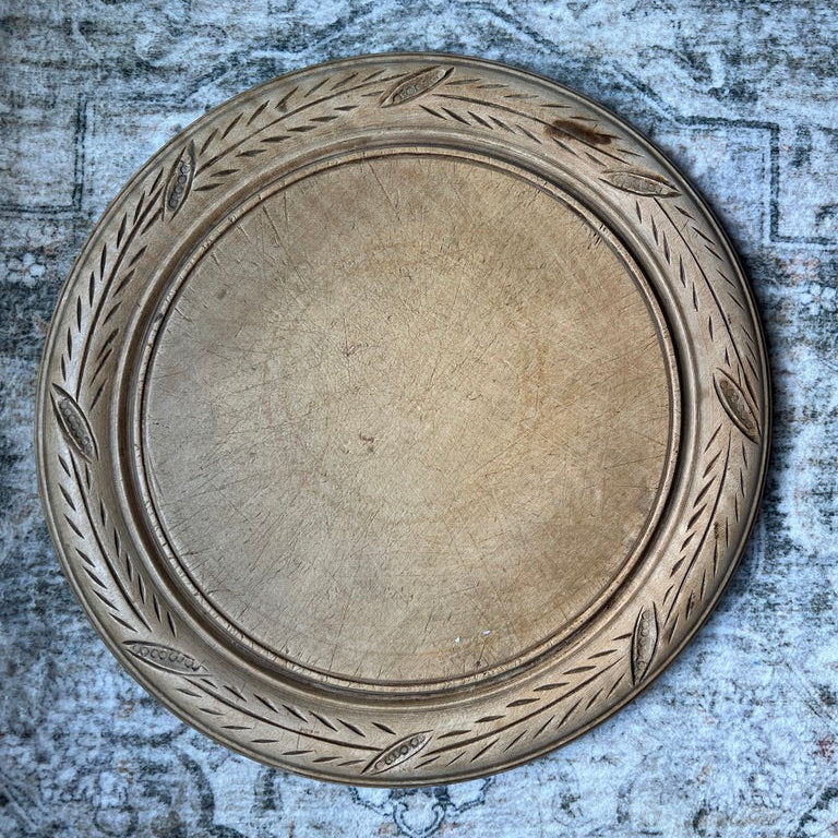 A round wooden serving tray with a carved leafy border, shown from a top-down view on a textured, distressed gray background.