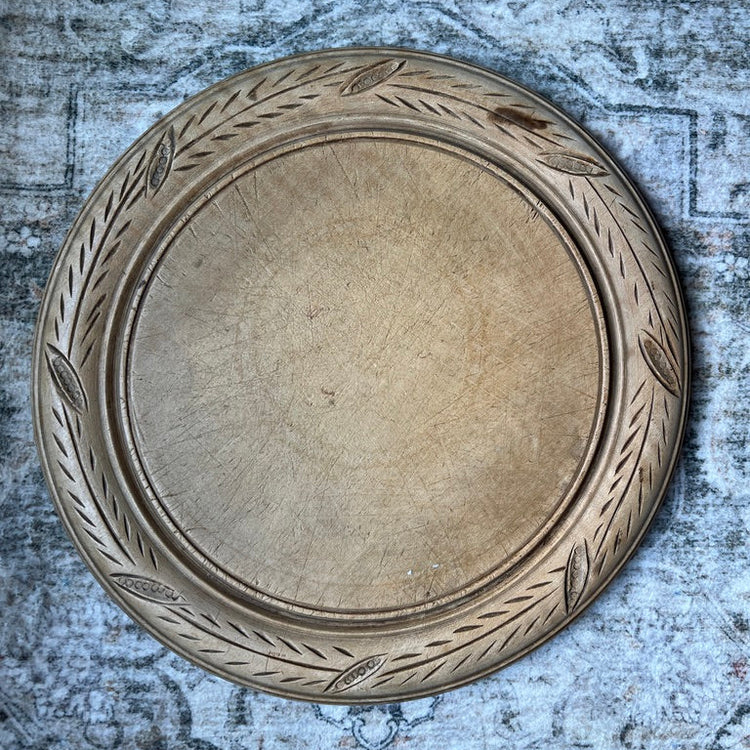 A round wooden serving tray with a carved leafy border, shown from a top-down view on a textured, distressed gray background.
