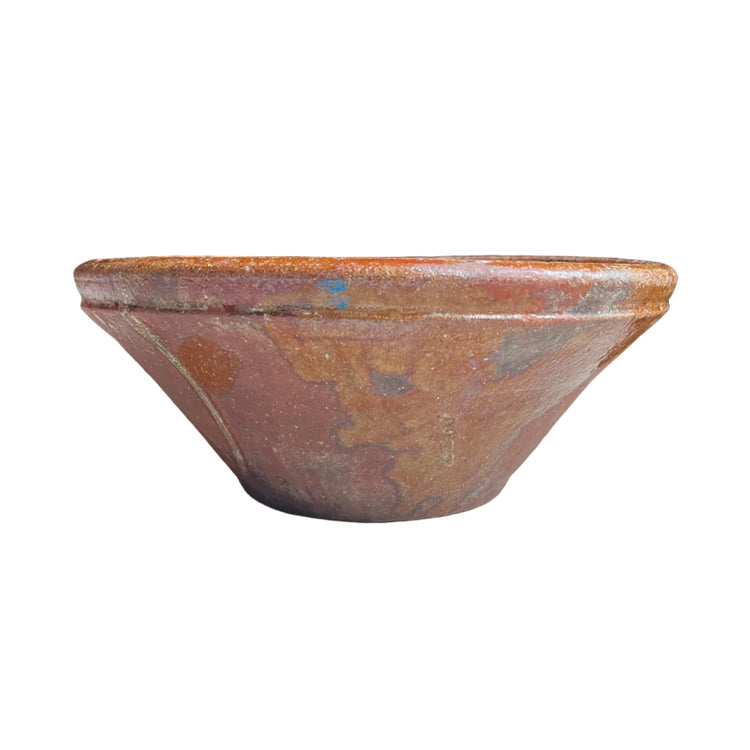 A rustic, hand-painted terracotta clay bowl with a wide rim, sitting on a light gray textured surface. The bowl features a mottled finish with earthy tones of terracotta, brown, and subtle hints of blue and gold.