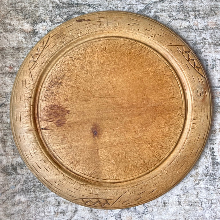 A round wooden serving tray with a carved geometric border, shown from a top-down view on a distressed gray patterned rug.