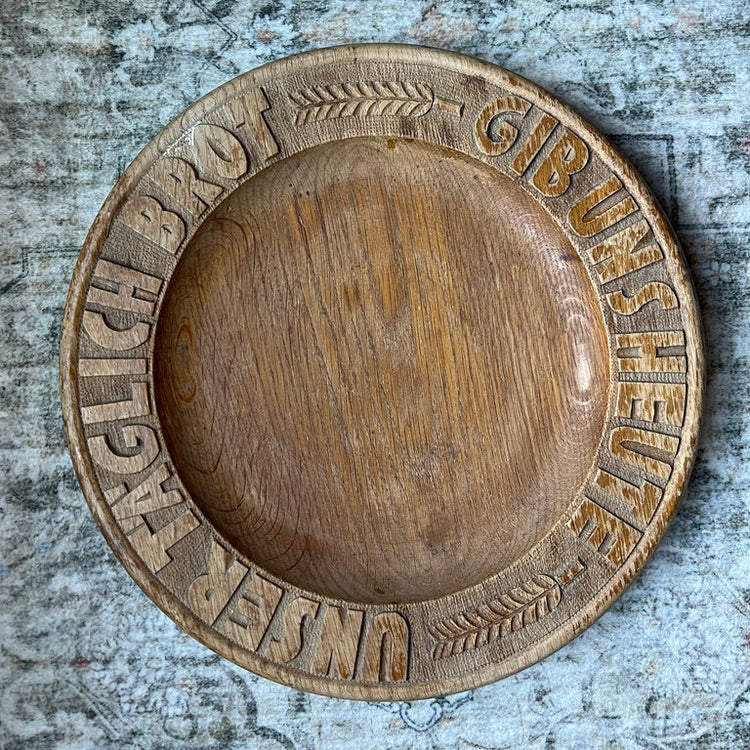 A round, hand-carved wooden serving platter with German text 'UNSER TÄGLICH BROT - GIB UNS HAUpte' carved around the rim, displayed on a textured, weathered gray-blue surface.