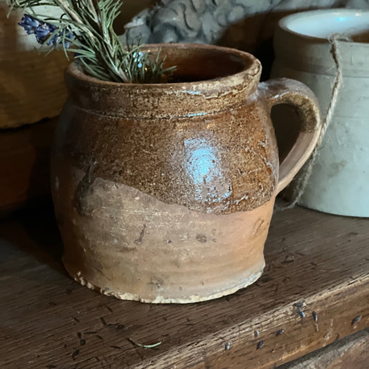 A rustic handmade ceramic pot with a handle, featuring a two-tone brown and beige glaze, holding a sprig of rosemary on a wooden surface.