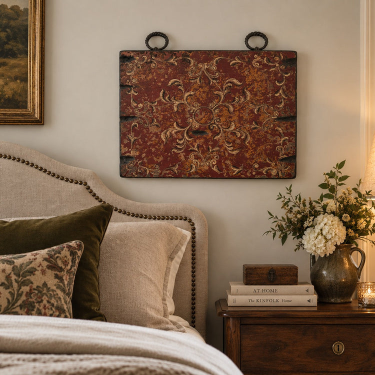 A rustic rectangular wall hanging with a distressed red and gold damask pattern, suspended above a bed in a warm, styled bedroom setting.