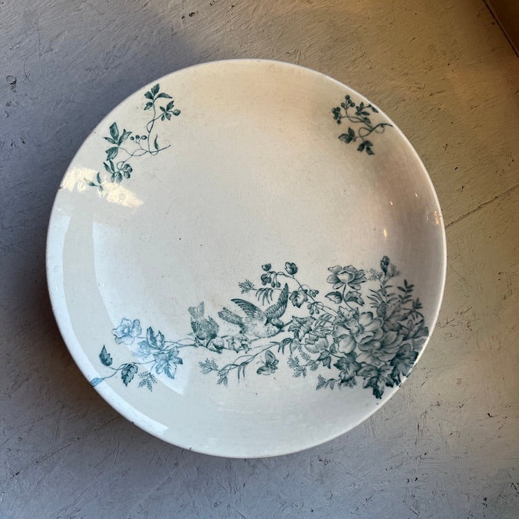 A round white ceramic plate with a delicate blue floral and leaf pattern, photographed from above on a textured gray surface.