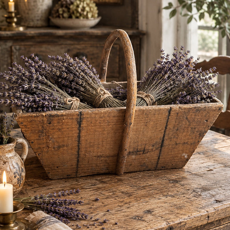 A rustic wooden basket with a single handle, filled with bundles of dried lavender, displayed on a weathered wooden table in a cozy, vintage-style room with a candle and plants.