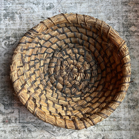 A round, hand-woven basket made from natural straw and dark brown accents, photographed from directly above on a distressed, vintage-style gray patterned rug.