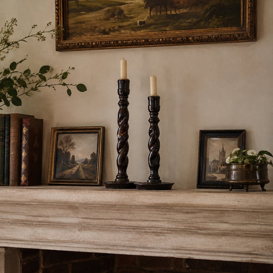 A styled interior scene showing a fireplace mantel with a large ceramic vase of greenery, books, two dark twisted candlesticks with white taper candles, small framed landscape paintings, and a potted plant against a warm beige wall.