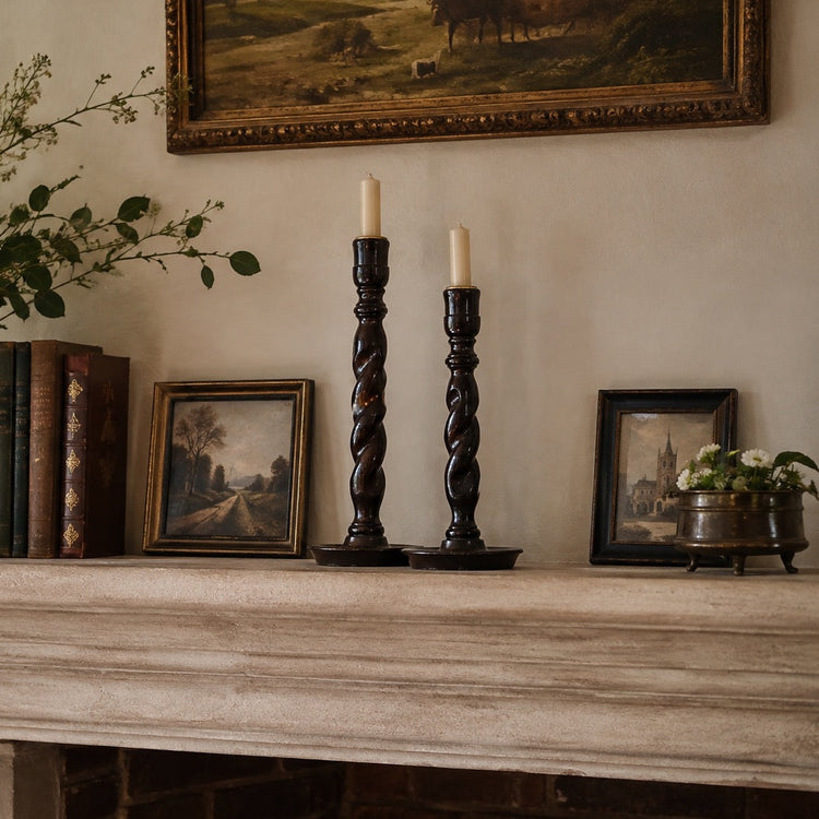 A styled interior scene showing a fireplace mantel with a large ceramic vase of greenery, books, two dark twisted candlesticks with white taper candles, small framed landscape paintings, and a potted plant against a warm beige wall.