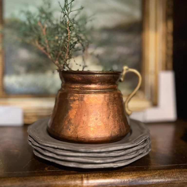 A distressed copper pitcher with a gold-toned handle, filled with greenery, sitting on a stack of gray ceramic plates on a dark wooden table with a blurred framed picture in the background.