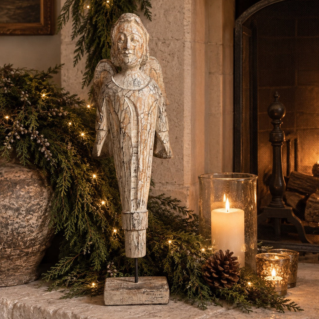 A weathered stone angel figurine stands on a fireplace mantel surrounded by green garlands, warm string lights, a lit pillar candle in a glass holder, and a small glowing candle in a metal container.