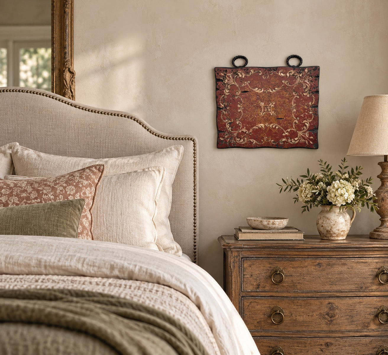 A rustic wall hanging with an ornate red and gold damask pattern is displayed above a wooden nightstand in a beautifully styled bedroom.