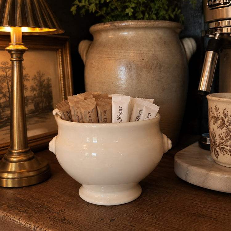 A cream-colored ceramic sugar bowl filled with sugar packets, set on a rustic wooden table next to a brass table lamp and a coffee machine.
