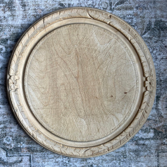 A round, carved wooden decorative tray or serving platter with an ornate raised border, shown from a top-down view on a textured gray rustic background.