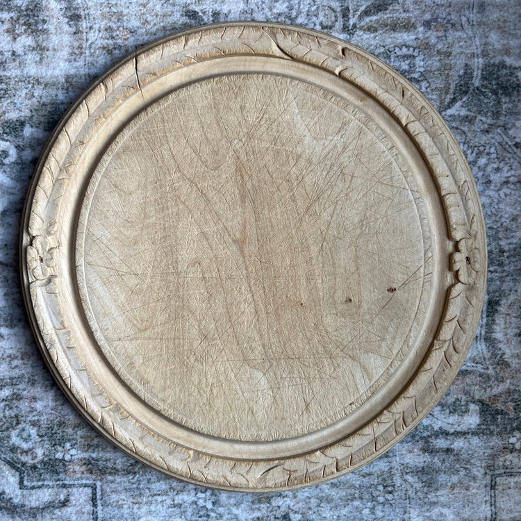 A round, carved wooden decorative tray or serving platter with an ornate raised border, shown from a top-down view on a textured gray rustic background.
